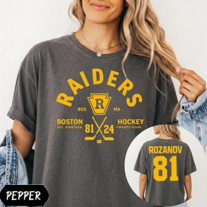 May include: A charcoal gray short-sleeve t-shirt featuring yellow graphics. The front displays the text RAIDERS, BOSTON HOCKEY, EST. NINETEEN 81 24, and a hockey emblem. The back shows the name ROZANOV and the number 81 in large yellow lettering.