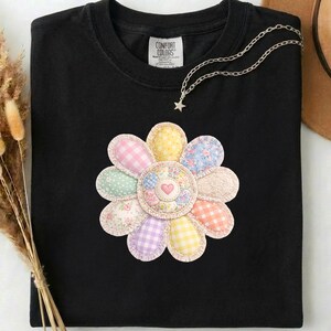 Puede incluir: Camiseta negra con un gran diseño floral colorido. La flor está hecha de varias telas estampadas, incluyendo cuadros y estampados florales, con un corazón en el centro. Un collar plateado con un colgante de estrella está sobre la camiseta.