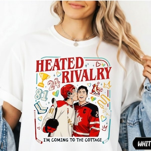May include: A white t-shirt featuring a graphic print with the text HEATED RIVALRY and I'M COMING TO THE COTTAGE. The design includes two hockey players, one kissing the other on the cheek, surrounded by hockey gear, a trophy, and colorful hearts.