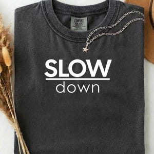 May include: Charcoal grey t-shirt featuring the white text "SLOW down". A silver necklace with a star charm is draped on the shirt. The t-shirt has a crew neck and a "Comfort Colors" label.