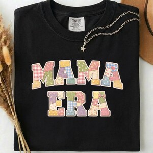 May include: Black t-shirt with the words "MAMA ERA" in a patchwork design. The letters are in various pastel colors and patterns. A silver necklace with a star pendant is draped on the shirt.