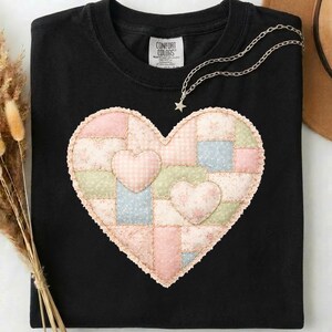 May include: Black t-shirt with a patchwork heart design in pastel pink, blue, and green. Two smaller hearts are appliquéd onto the larger one. A silver necklace with a star pendant is draped on the shirt.