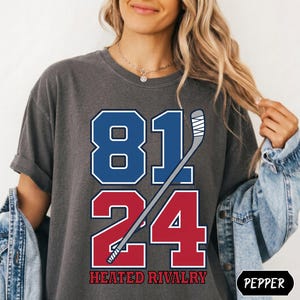May include: A charcoal gray short-sleeve t-shirt featuring a graphic print with the blue number 81 above the red number 24. A white hockey stick is positioned diagonally across the numbers. The text HEATED RIVALRY is printed in red below.