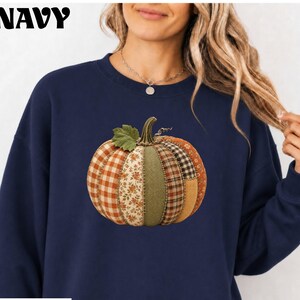 May include: A navy blue crewneck sweatshirt featuring a large, central graphic of a pumpkin. The pumpkin is composed of vertical segments with various patterns, including orange gingham, floral print, solid olive green, and brown plaid fabric.