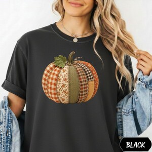 Op de afbeelding: Een zwart t-shirt met een centrale afbeelding van een pompoen die is opgebouwd uit verschillende stofachtige segmenten, waaronder oranje-witte vichy-ruitjes, bloemmotieven, effen olijfgroen en bruin ruitpatroon, bekroond met een groen blad.