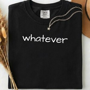 Op de afbeelding: Zwart T-shirt met het woord "whatever" in het wit, een zilveren ketting met een sterhanger en een Comfort Colors-label. Het shirt is netjes opgevouwen, met een neutrale achtergrond en gedroogde planten.