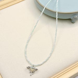 May include: A necklace featuring a strand of small, pale iridescent beads and a silver-toned pendant. The pendant consists of a heart shape encircled by a ring, with metallic drip-like accents extending from the bottom of the heart structure.