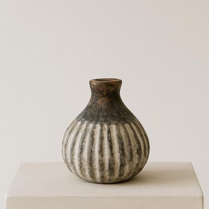 Może przedstawiać: Ręcznie wykonany ceramiczny wazon o pękatym korpusie i wąskiej szyi. Wazon posiada pionowe żebrowane detale i dwutonową kolorystykę, z ciemnobrązową górą i jasnoszarym i białym korpusem. Wazon jest eksponowany na cokole.