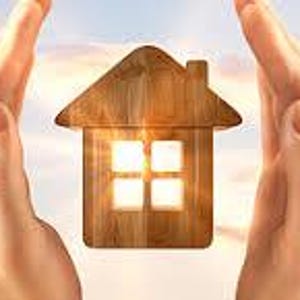May include: A wooden house cutout with four square windows is held between two hands. The house has a pitched roof and a chimney. The background is a sky with clouds and sunlight.