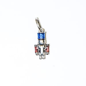May include: A silver-toned charm in the shape of a toy soldier nutcracker. The nutcracker has a blue hat, red arms, and is detailed with white and silver accents. The charm is from the Pandora brand.
