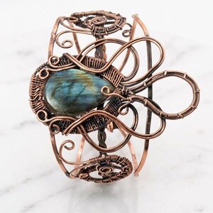 May include: A copper wire-wrapped cuff bracelet featuring a central teardrop-shaped labradorite gemstone with blue and green iridescence. The metalwork consists of intricate swirls, loops, and coiled wire patterns that form an open, airy structure.