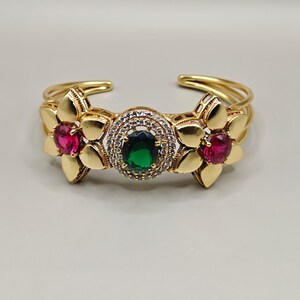 Peut inclure: Bracelet manchette doré orné de motifs floraux. Le bracelet présente une pierre verte centrale entourée de cristaux transparents, flanquée de deux pierres roses. Les éléments floraux sont détaillés.