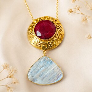 May include: A gold-tone necklace featuring a circular pendant with an intricate scrollwork pattern and a faceted deep red gemstone at its centre. A teardrop-shaped, pale blue, translucent stone hangs from the bottom of the circular pendant.