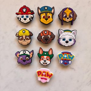 May include: A selection of colourful cartoon dog face charms, featuring characters in hats and helmets. The charms include the Paw Patrol logo and various dog breeds. The charms are made of a glossy material.