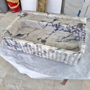 Peut inclure: Évier rectangulaire en marbre avec un motif marbré blanc et gris et des veines violet foncé. L'évier a un bord cannelé et un seul trou d'évacuation. Il repose sur une feuille de papier.