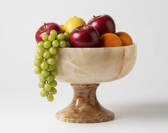 May include: A decorative fruit bowl filled with fresh fruit. The bowl is made of a light-colored, marbled material and sits on a pedestal base. The bowl contains red apples, green grapes, oranges, and a yellow lemon.