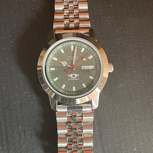 May include: A silver-toned Citizen automatic watch with a green dial and a stainless steel band. The watch face displays the day and date, with red and white accents. The watch band is made of multiple small, connected links.