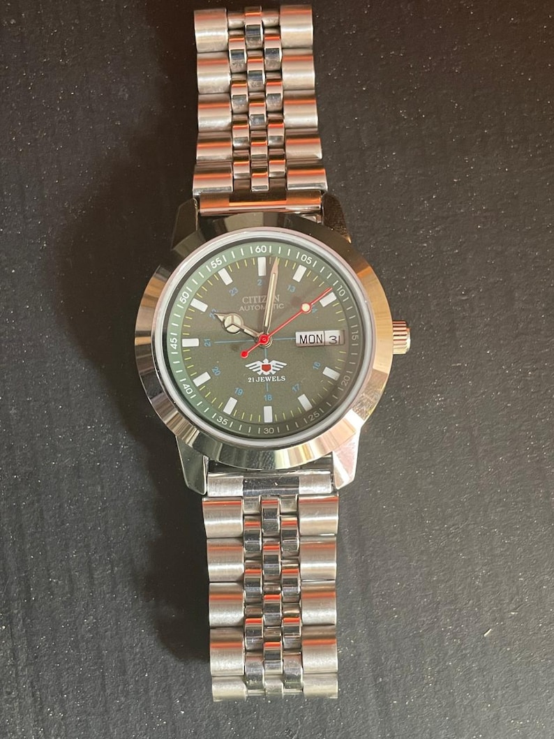 May include: A silver-toned Citizen automatic watch with a green dial and a stainless steel band. The watch face displays the day and date, with red and white accents. The watch band is made of multiple small, connected links.