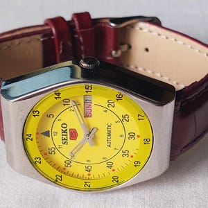 Puede incluir: Un reloj automático Seiko vintage con esfera amarilla y caja plateada. El reloj tiene una correa de cuero burdeos. La esfera muestra el día y la fecha, con el logotipo de Seiko en un lugar destacado.