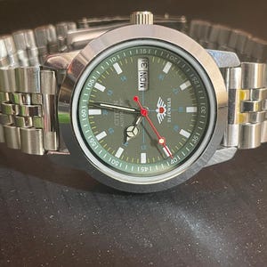 May include: A silver-toned Citizen automatic wristwatch with a green face and a stainless steel band. The watch face displays the day and date, with red and white accents. The watch is on a dark surface.