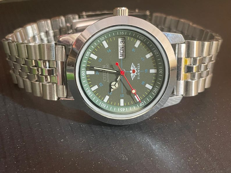 May include: A silver-toned Citizen automatic wristwatch with a green face and a stainless steel band. The watch face displays the day and date, with red and white accents. The watch is on a dark surface.