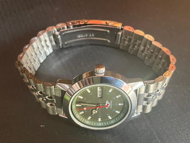 May include: A stainless steel wristwatch with a silver band and a dark green face. The watch face displays the day of the week and the time. The watch band is made of small, connected metal links.