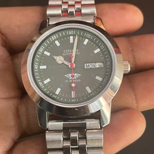 May include: A silver Citizen automatic wristwatch with a dark green face. The watch features a day and date display, a red second hand, and a silver band. The watch face displays the text "CITIZEN AUTOMATIC" and "21 JEWELS".