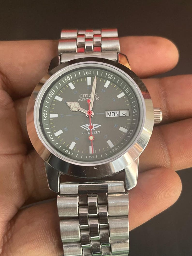 May include: A silver Citizen automatic wristwatch with a dark green face. The watch features a day and date display, a red second hand, and a silver band. The watch face displays the text "CITIZEN AUTOMATIC" and "21 JEWELS".