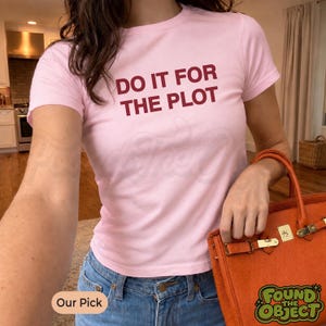 Könnte beinhalten: Ein hellrosa kurzärmeliges T-Shirt mit dem Text DO IT FOR THE PLOT in dunkelroten Buchstaben auf der Brust. Das T-Shirt wird mit einer blauen Jeans und einer orangefarbenen Handtasche mit goldfarbenen Metallbeschlägen und Verschluss kombiniert.