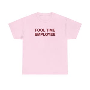 Op de afbeelding: Een lichtroze T-shirt met korte mouwen en de tekst FOOL TIME EMPLOYEE in donkerrode hoofdletters op het midden van de borst. Het kledingstuk heeft een ronde hals en een standaard pasvorm met een effen ontwerp aan de voor- en achterkant.