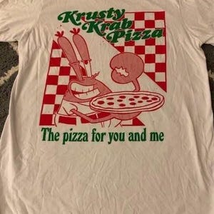 Pode incluir: Uma t-shirt branca com um gráfico vermelho e verde do Sr. Krabs a segurar uma pizza. O design inclui um fundo axadrezado vermelho e branco com o texto Krusty Krab Pizza e The pizza for you and me impresso em letras verdes.