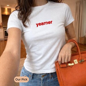 Puede incluir: Una camiseta blanca de manga corta con la palabra "yearner" impresa en letras minúsculas rojas en el pecho. La prenda se combina con unos vaqueros azules y un bolso de mano naranja texturizado con hebillas y herrajes de metal dorado.
