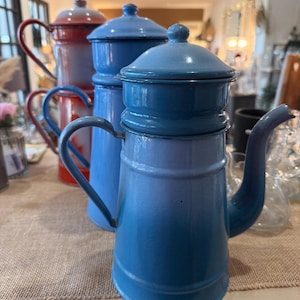 Puede incluir: Tres cafeteras vintage de esmalte en tonos azules y rojos. Las cafeteras tienen boquillas curvas, asas y tapas abovedadas. Están dispuestas sobre una superficie clara, con una tela de tono neutro debajo.