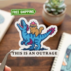 May include: A sticker featuring a blue dinosaur with pink accents and a brown cat wearing sunglasses and a crown. The text "THIS IS AN OUTRAGE" is below the image. The sticker is on a wooden surface.