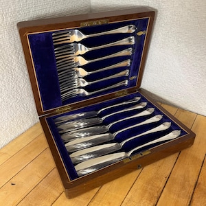 Peut inclure: Un coffret en bois à couvercle articulé, doublé de velours bleu, contient un ensemble de couverts en argent. L'ensemble comprend des fourchettes et des couteaux, tous avec une finition argentée brillante. La boîte est ouverte, présentant les couverts.
