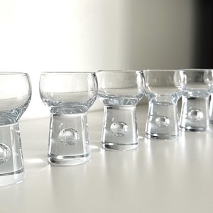 May include: A set of six clear glass shot glasses. Each glass features a rounded bowl and a base with a spherical detail. The glasses are arranged in a row on a white surface, highlighting their elegant design. A stylish addition.