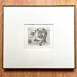 Peut inclure: Une gravure encadrée en noir et blanc d'un homme avec des poissons nageant autour de son visage. L'œuvre est encadrée en blanc et dans un cadre doré. L'image est sur une surface en bois.
