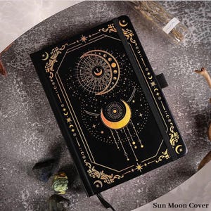 May include: Black journal with a celestial sun and moon design in gold. The cover features intricate details, including stars and crescent moons. The text "Sun Moon Cover" is visible on the bottom of the image. A black elastic band and ribbon bookmark are included.