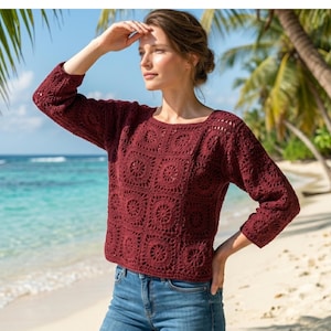 May include: A burgundy crochet sweater with a square pattern and long sleeves. The sweater is paired with blue jeans. The image is taken on a beach with palm trees and the ocean in the background.