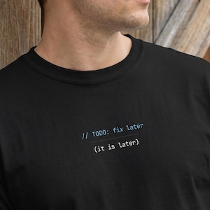 May include: Black t-shirt with the text "// TODO: fix later (it is later)" in light blue. The shirt is made of a soft material. A person is holding a coffee cup.