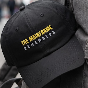 May include: A black baseball cap featuring the text THE MAINFRAME REMEMBERS embroidered in yellow and white on the front panel. The cap has a structured crown, a curved brim, and a button on top, resting against a grey canvas backpack.
