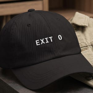 May include: A black baseball cap featuring the white embroidered text EXIT 0 on the front panel. The cap has a structured crown, a curved brim, and a button on top. The design is simple and clean, showcasing the white lettering against the black fabric.