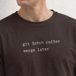 Op de afbeelding: Een donkerbruin t-shirt met de tekst "git fetch coffee merge later" in het wit. Het shirt is een T-shirt met korte mouwen en een ronde hals.