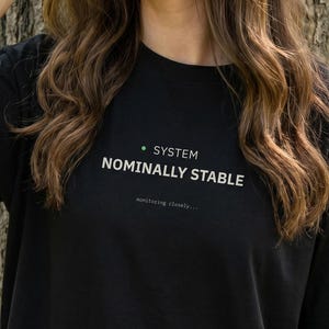 May include: Black t-shirt with the text "SYSTEM NOMINALLY STABLE" in white, with a green dot above the word "SYSTEM". The shirt is being worn outdoors near a tree.