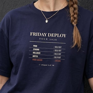 Puede incluir: Una camiseta de manga corta azul marino con texto en blanco y rojo que dice FRIDAY DEPLOY TOUR 2026, con una lista de elementos que incluye PROD, STAGING, ROLLBACK, HOTFIX y PROD AGAIN, marcados como SOLD OUT o ENCORE, y texto pequeño.