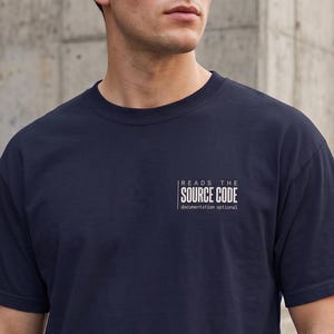 May include: A navy blue short-sleeved t-shirt featuring white text on the left chest that reads "READS THE SOURCE CODE" with the smaller text "documentation optional" underneath, all enclosed within a thin white rectangular border.