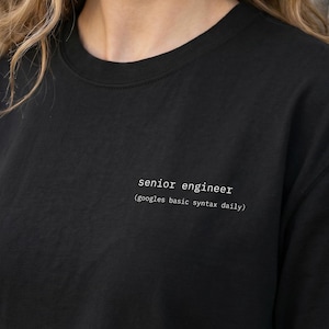May include: Black crew neck t-shirt with the text "senior engineer (googles basic syntax daily)" printed in white. The shirt is made of a soft material and has short sleeves.