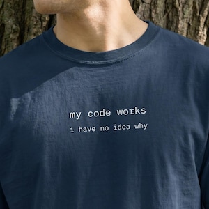 Puede incluir: Camiseta azul marino con el texto blanco "my code works, i have no idea why". La camiseta es de cuello redondo y parece estar hecha de un material suave. La imagen está tomada al aire libre con un árbol de fondo.