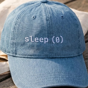 May include: A light blue denim baseball cap with the word "sleep (0)" embroidered in white. The cap is on a wooden surface next to a beige tote bag.
