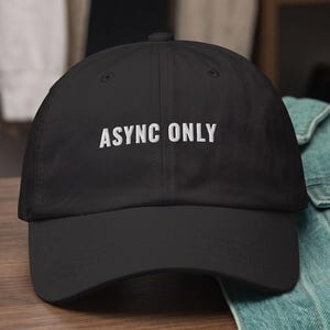 Puede incluir: Una gorra de béisbol negra con el texto bordado en blanco ASYNC ONLY centrado en el panel frontal. La gorra tiene una corona estructurada, una visera curva y un diseño clásico de seis paneles con ojales de ventilación en cada panel.